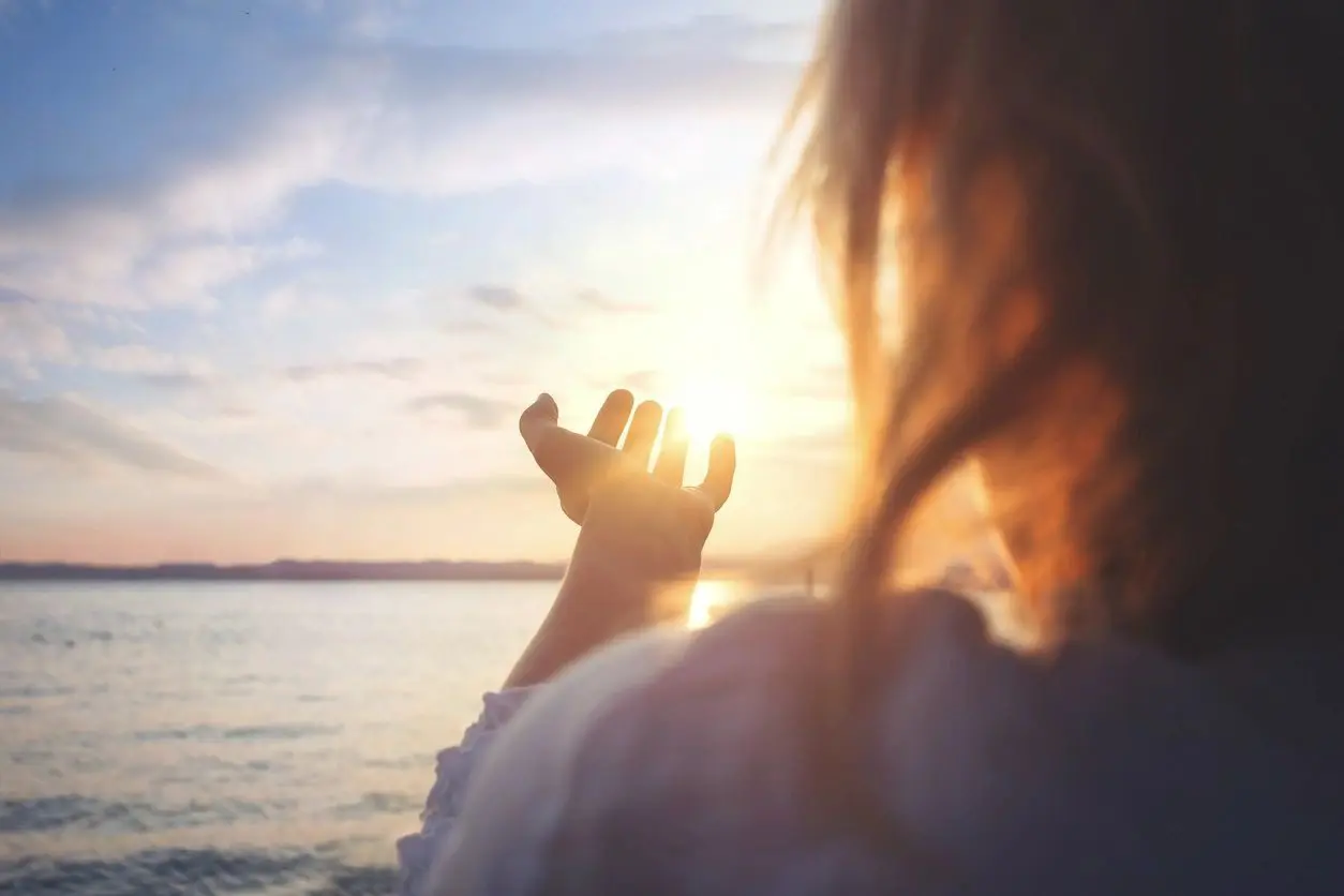 A person faces a body of water at sunset, extending a hand toward the sun, as though channeling the energy from an enlightening tarot reading class.