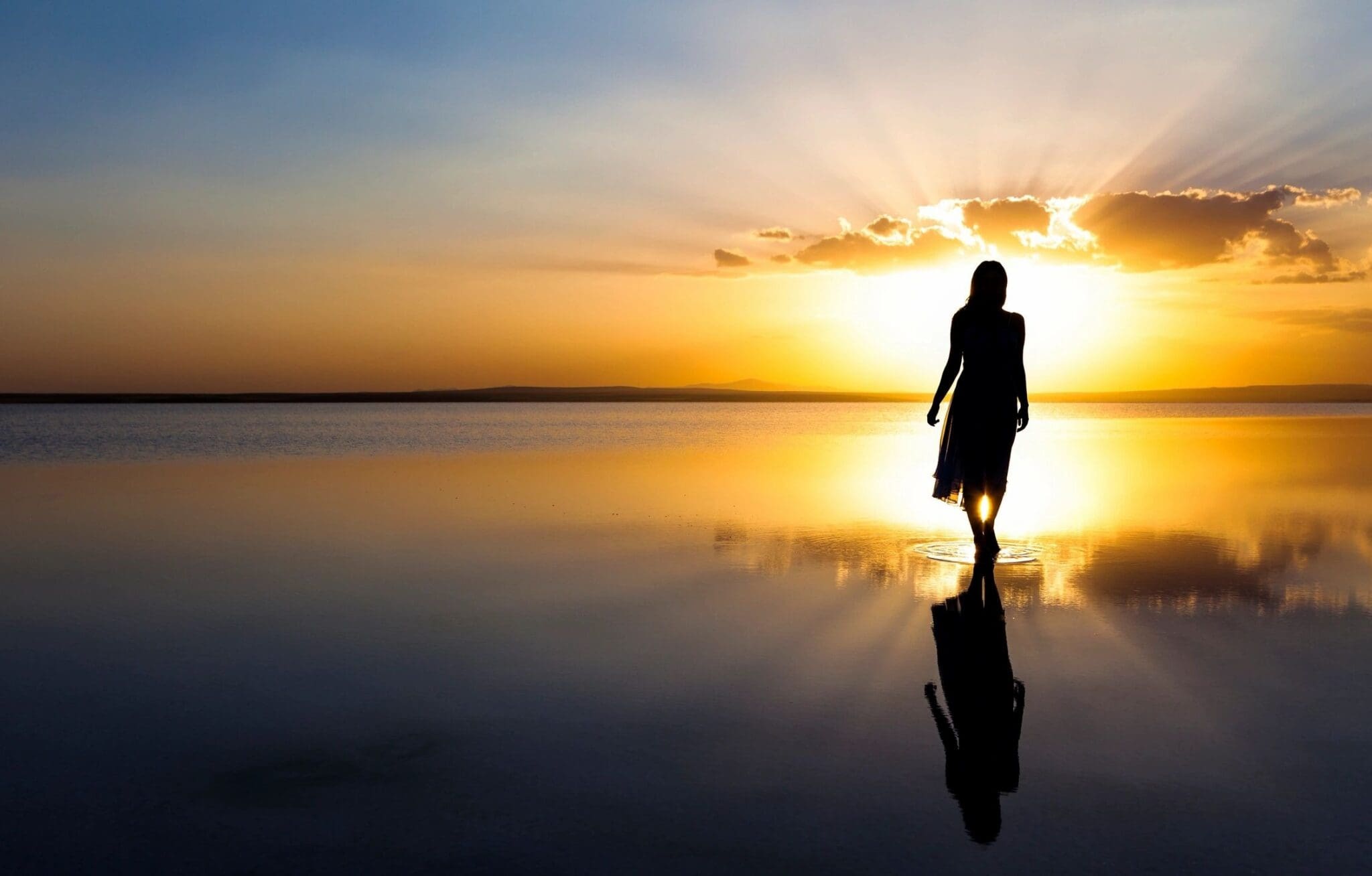 Silhouette of a person walking on a calm, reflective water surface at sunset with a clear sky and minimal clouds in the background.