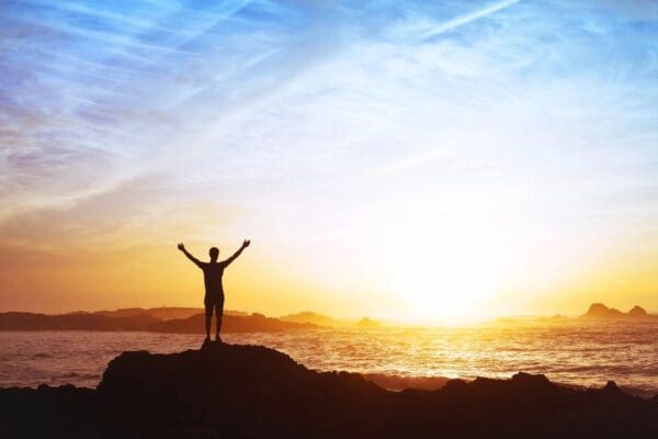 Silhouette of a person standing on a rocky outcrop with arms raised, facing a sunrise or sunset over the ocean, as if drawing energy akin to psychic tarot readings from the horizon's mystical glow.