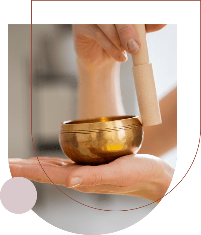 A person's hands holding a small brass singing bowl and wooden striker, positioned against a blurred background, reminiscent of the serene ambiance often found in Tarot reading classes.