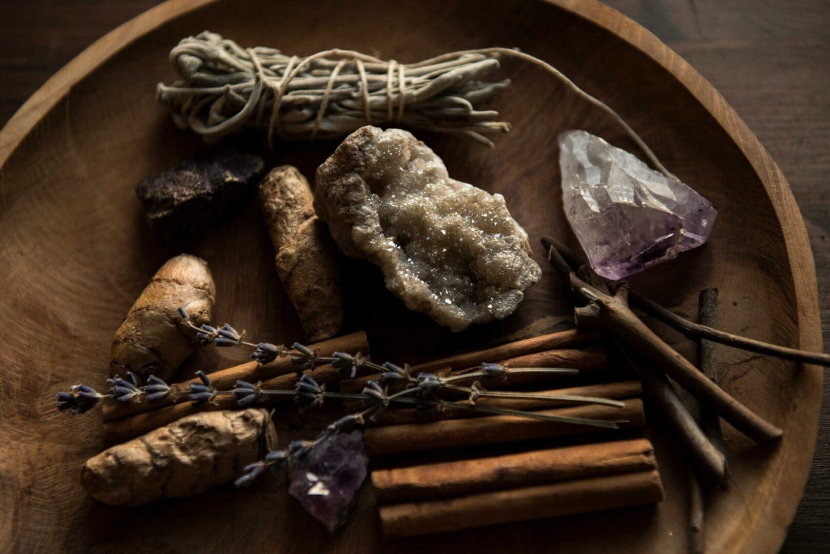 A wooden bowl contains sage, various crystals, pieces of ginger, cinnamon sticks, and dried lavender.