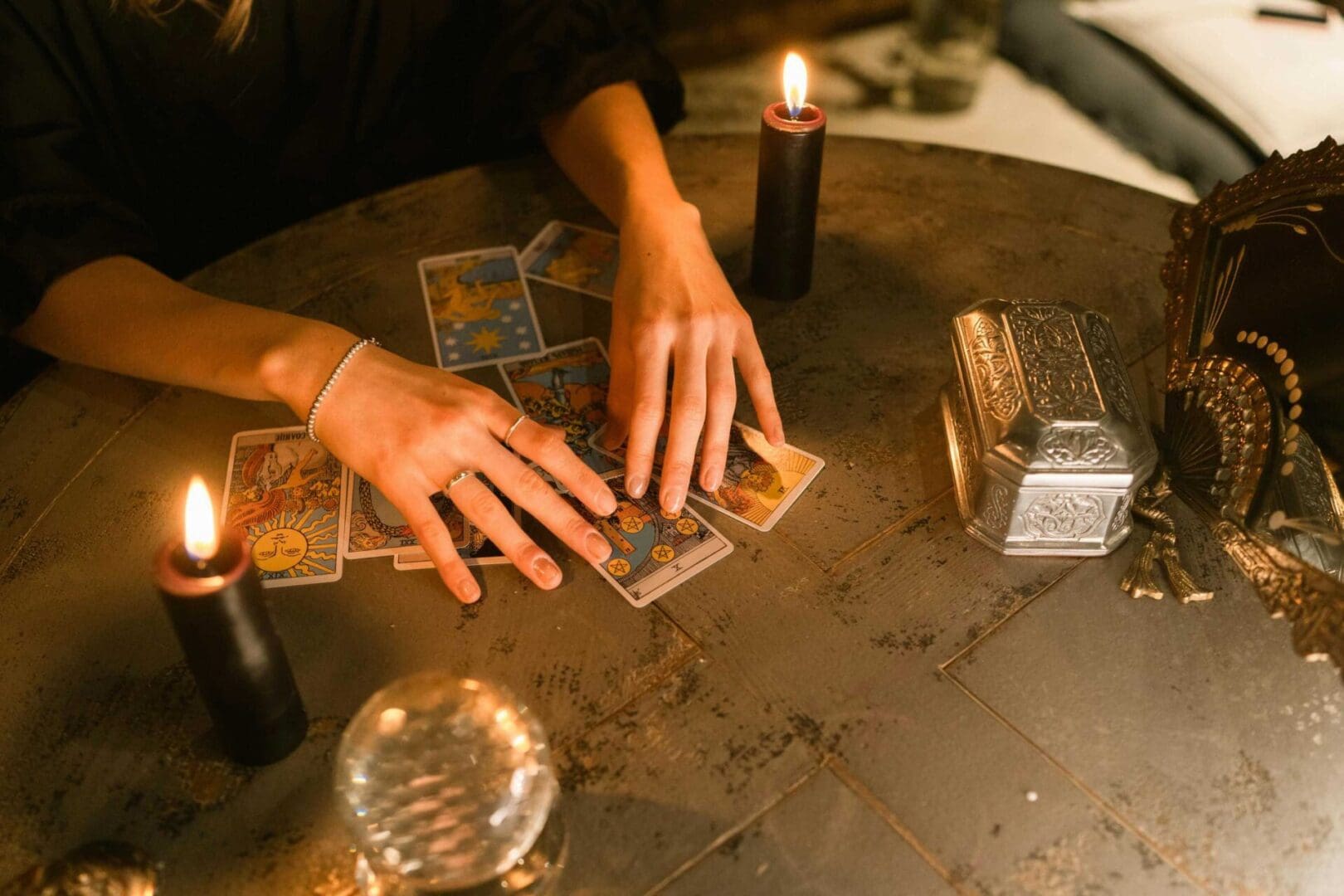 A person arranges tarot cards on a table surrounded by lit candles, a crystal ball, and a decorative box.