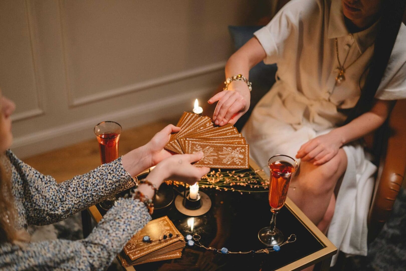 Two individuals are sitting at a table with lit candles and two glasses of red liquid. One person is holding a fanned-out set of tarot cards, handing them to the other person.