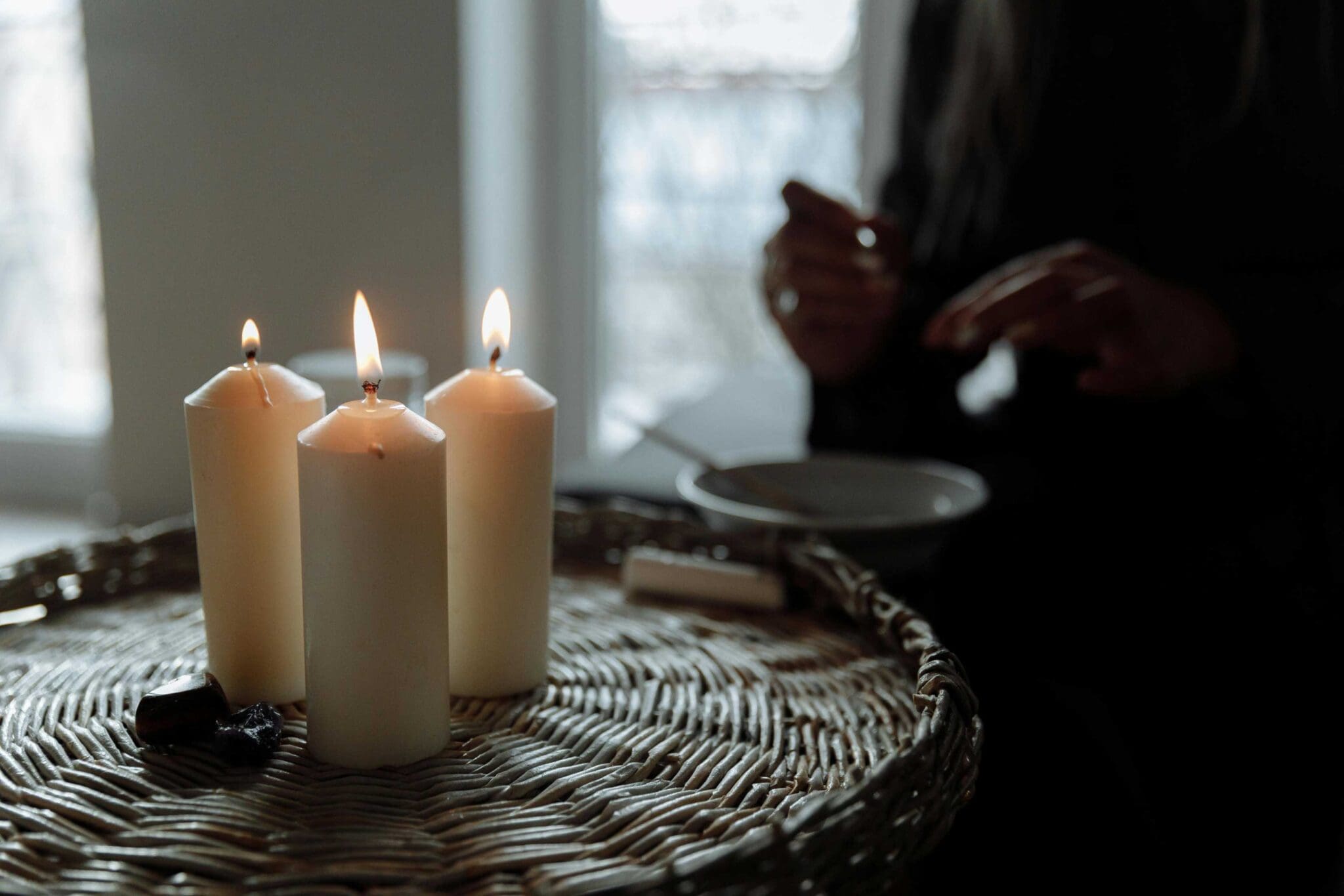 Three lit candles are placed on a wicker surface in the foreground, while a person with partially visible hands sits in the dimly lit background.