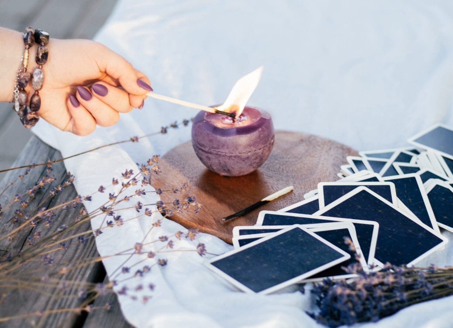 Woman's hand in bracelet with purple long nails lights candle with match, next to deck of cards and lavender flowers on white and wooden surface