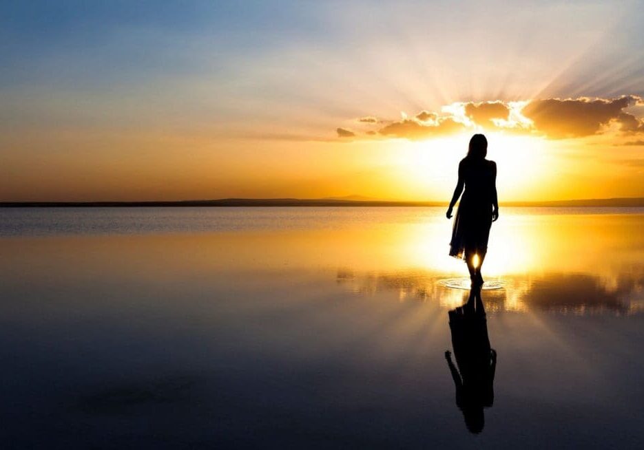 Silhouette of a person walking on a calm, reflective water surface at sunset with a clear sky and minimal clouds in the background.