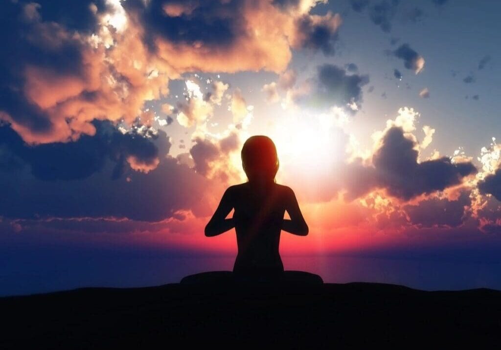 A silhouette of a person meditating outdoors against a backdrop of a colorful sunset sky with clouds.