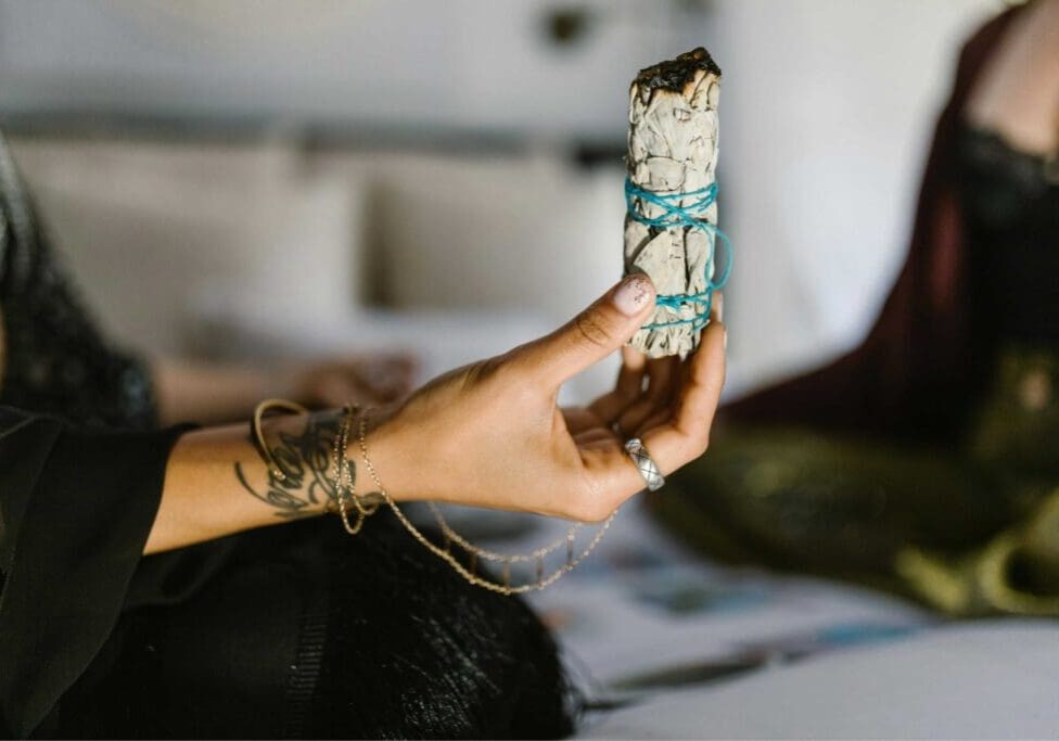 A person holds a bundle of sage wrapped in blue string, with visible tattoos and rings on their hand.