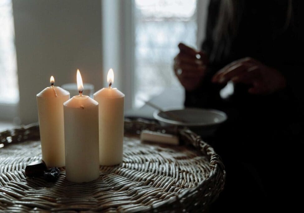 Three lit candles are placed on a wicker surface in the foreground, while a person with partially visible hands sits in the dimly lit background.