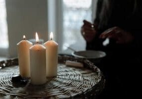 Three lit candles are placed on a wicker surface in the foreground, while a person with partially visible hands sits in the dimly lit background.